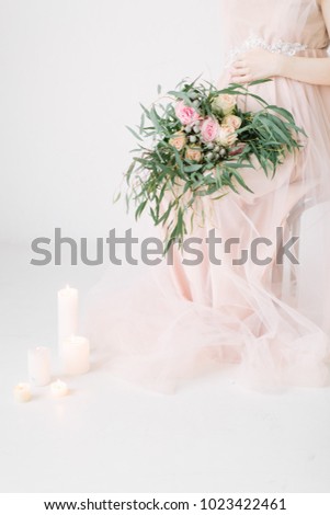 Pregnant girl sits on a decorated chair with large bouquet of flowers with candles in the ground on white background