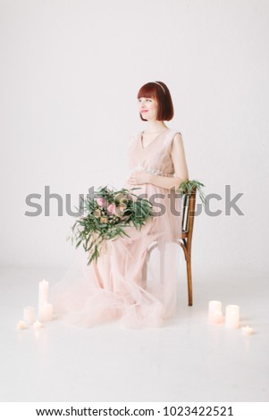 Pregnant girl sits on a decorated chair with large bouquet of flowers with candles in the ground on white background