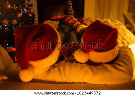 Couple wearing Santa hats, drinking mulled wine, sitting in woolen socks near fireplace in living room decorated for Christmas holidays, hugging and enjoying life