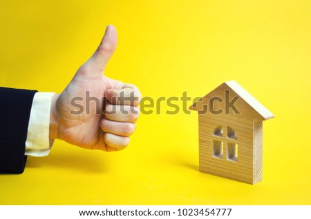 Finger up and house. Wooden house and approving hand. A businessman in a suit gives a good appraisal to the business. Real estate, good make, excellent price. Yellow background.