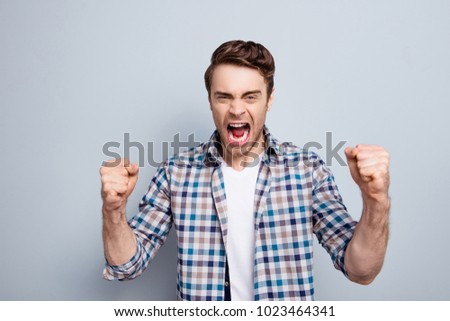 Aggressive man in checkered shirt with raised fists and open mouth is out of himself, yelling, screaming, shouting with cruelty over grey background