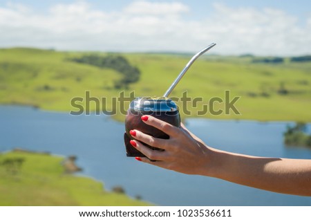 Hand of woman holding traditional mate, mate, with green field view. Royalty-Free Stock Photo #1023536611