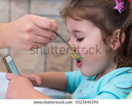 Mom feeding cute little girl with soup while watching cartoons on mobile phone