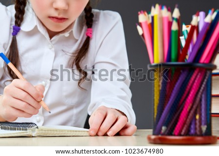Back focus with shallow depth of field picture of smiling beautiful cute little girlwriting something on the grey background. Her hair is done in plaits. There are some pencils in front of the girl.
