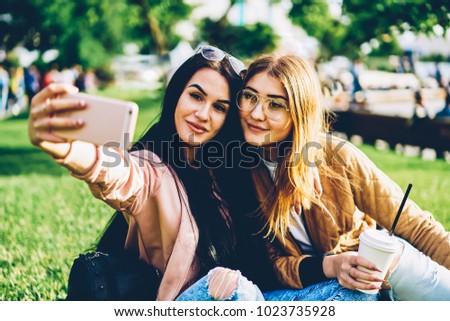 Cheerful female friends making beautiful pictures on smartphone using application on smartphone.Attractive young women making selfie on modern mobile posing outdoors during free time on weekend