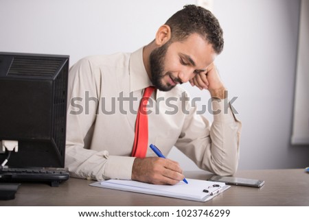 A young employee sitting on his desk writing on the clipboard focus, putting his hand on cheek,