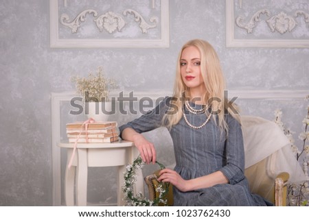 The blonde women in a gray dress and beads around her neck sits on a chair next to her and put her hand on a vintage white table with books in the interiors