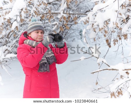Merry rosy-cheeked girl in a red jacket and light knit hat, scarf and gloves pictures of winter forest after a snowfall and keep the camera in landscape position