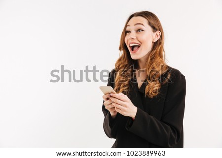 Beautiful curly woman in black jacket looking aside with surprise while holding and using cell phone isolated over white background