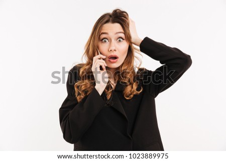 Picture of attractive woman with long curly hair in black outfit being surprised and grabbing head while having mobile call isolated over white background