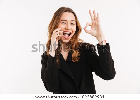 Cheery businesslike woman with long auburn hair in black jacket talking on smartphone and gesturing ok sign isolated over white background