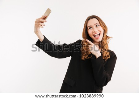 Photo of amusing young woman in black jacket making selfie with smile and pointing finger on mobile phone isolated over white background