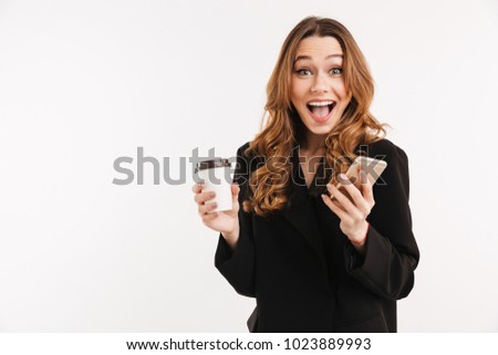Long-haired brunette woman in black outfit smiling on camera while holding smartphone and takeaway coffee in hands isolated over white background