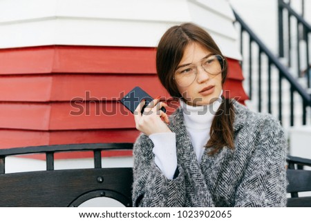 beautiful stylish girl in trendy glasses and a gray coat is sitting on the street, holding a smartphone in her hand