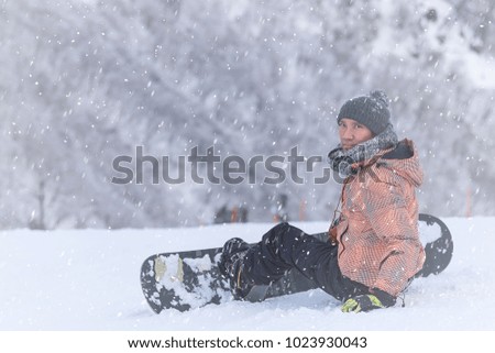 Portrait of snowboarder wearing sweater hat, orange & black stripe jacket, gloves and pants sitting on top of a mountain and looking at the camera on the background of hills. Snowflake, winter season