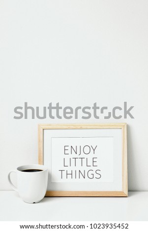 a wooden-framed picture with the text enjoy little things written in it and a cup of coffee, on a whit table against a white background with some blank space on top
