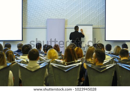 Speaker back to audience is going to write on board. Business meeting. Audience in the conference hall. Screens on the sides are isolated. Photo.