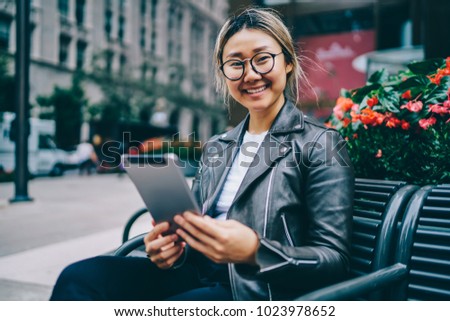 Portrait of cheerful Asian female in eyewear resting on bench on square blogging in social networks via digital tablet, Happy asian hipster girl using portable pc for chatting with friend outdoors