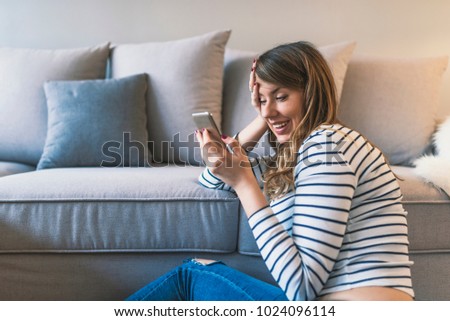 Lovely message from him. Attractive young woman looking at her smart phone and smiling while sitting on the carpet at home. Smiling young woman at home relaxing, she is using a smartphone and texting,