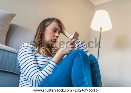 Portrait of young sad annoyed woman, receiving bad sms. Front view of a sad woman checking phone sitting on the floor in the living room at home. Sad news. Upset young woman with mobile phone 