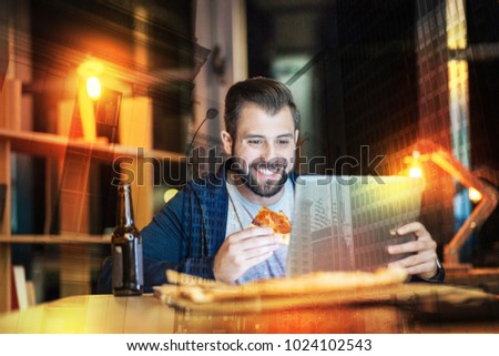 Eating alone. Cheerful emotional bearded man smiling and feeling happy while sitting at the table with a piece of pizza in his hand and looking at the screen of his convenient laptop