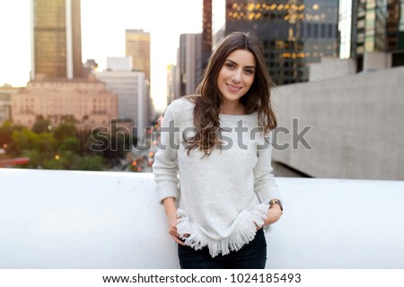 Young woman looking beautiful, sitting on a bridge across the boulevard in urban scenery, downtown, at sunset, smiling at camera.