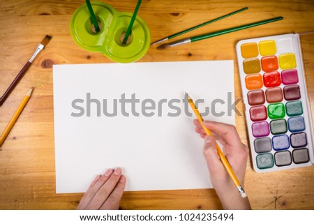 kids hand holding a pencil on blank page at the table with palette, empty space for text, top view
