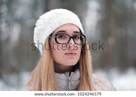 Portrait of a beautiful young lady on a winter forest background. Closeup face of the young lady with glasses and a white hat. In the background is blurred winter forest.