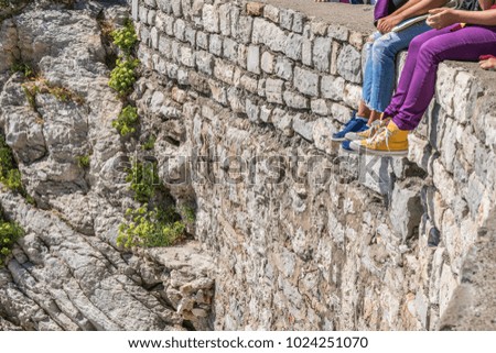Two persons are sitting on the stone wall near the beach in Budva, Montenegro. Purple and blue pants in the sunny day.  Royalty-Free Stock Photo #1024251070