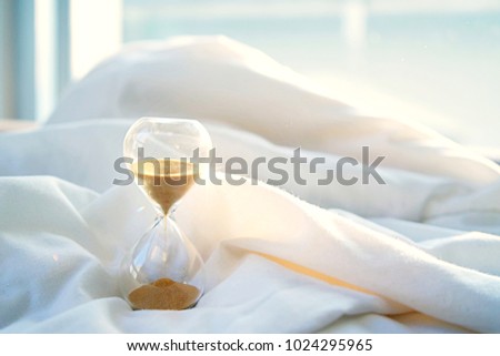 Lazy morning. Time passing concept. Hourglass on white blanket with morning light.
