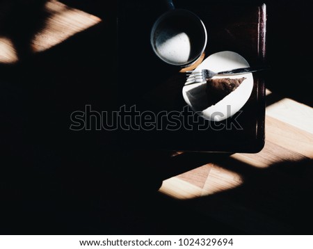 Top view picture of a white cup of cappuccino and chocolate cake on white plate above wooden table with window light effect.