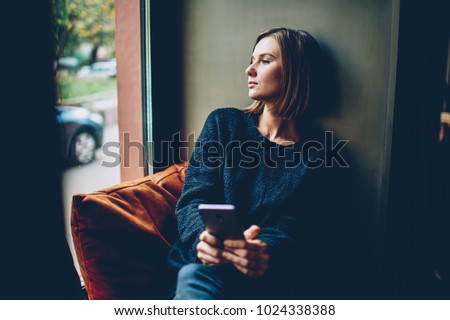 Thoughtful young woman dressed in black casual outfit looking out of window resting in comfortable coworking space. Pondering hipster girl with smartphone in hands thinking on future Royalty-Free Stock Photo #1024338388