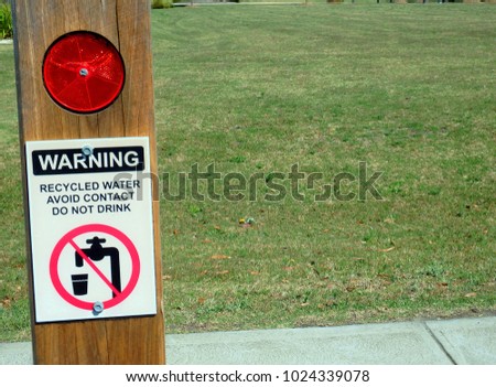 Warning recycled water avoid contact do not drink, sign in a timber pole with lawn in the background  placed near a side walk footpath.