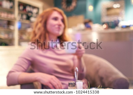 Blurred picture in the cafe. A young woman in a pink blouse with curly red hair sitting in a comfortable chair in a restaurant with a beautiful interior drinking coffee and eating dessert   
         