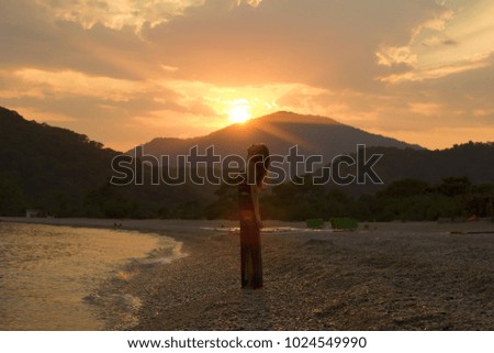 Girl stands on the sea beach. Beautiful sunset at the seaside with mountains on background. Sun breaks through the clouds. A stream of light in evening sky. Gorgeous woman silhouette. Ocean beach