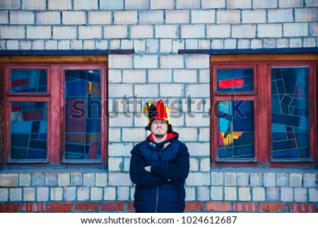 bearded brutal man in the hat of a buffoon stands near the brick wall and color windows