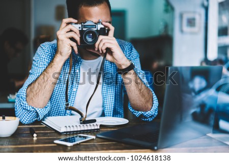 Skilled male photographer making picture of cafe sitting at table with netbook for editing,hipster guy enjoying photography spending time on hobby resting at coffee shop taking image on vintage camera