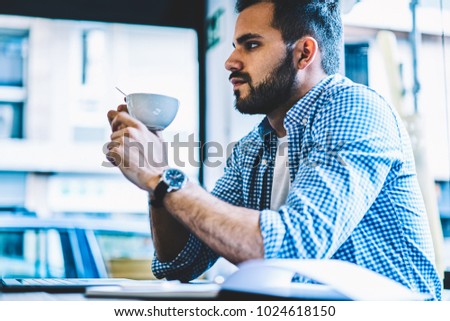 Serious young man concentrated on idea for online job during coffee break, thoughtful male it developer holding cup on free time sitting at working space with laptop computer in coworking office