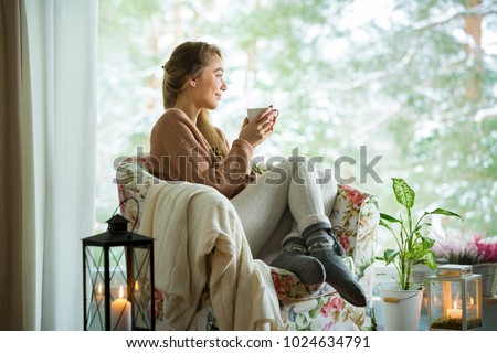 Young woman sitting home in a chair by the window with cup of hot coffee wearing knitted warm sweater. Cozy room decorated with lanterns and candles. Scenic view of pine trees in snow in window Royalty-Free Stock Photo #1024634791