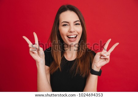 Portrait of adult brunette woman looking at camera with smile and showing victory sign with fingers isolated over red background
