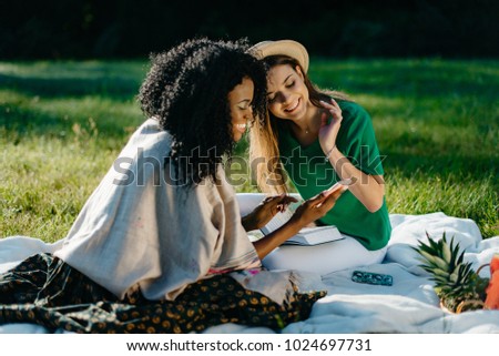 Outdoor portrait of two multi-race happy girl friends watching something on the mobile phone uring their picnic on the meadow.