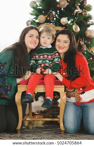 Two women hug a little boy while he sits on the chair before a Christmas tree