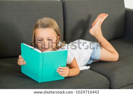 young sweet and happy little girl 6 or 7 years old lying on home living room sofa couch reading a book quiet and adorable in children education and lifestyle concept