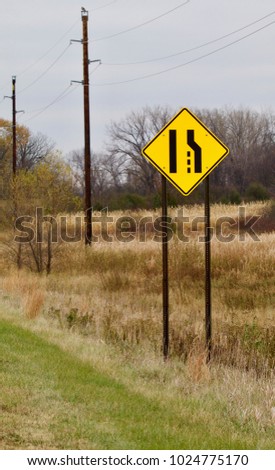 A bright yellow merging lanes warning sign shows against a rural backgound.