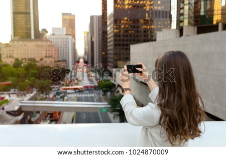 Beautiful young woman sitting on a bridge across the boulevard in urban scenery, downtown, at sunset, taking photos with smartphone.