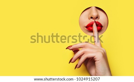 Red lips look through the hole in the yellow paper. A finger with a red varnish at the mouth.Fashion, beauty, make-up, cosmetics, beauty salon, style, personal care, geometry, texture, bright.