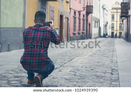 Back of man with red checkers shirt crouching to take photo with phone of empty street in old town Las Palmas, Gran Canaria. Young tourist visiting colorful buildings street in Canary Islands