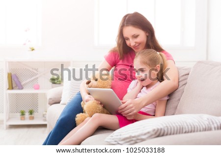 Happy girl and her mom using digital tablet, sitting on sofa at home. Mother showing her daughter photos, cartoon, playing developing games, copy space