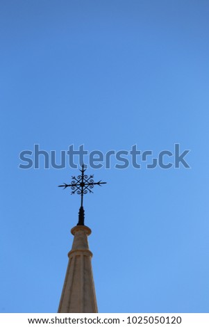 Outdoor view of a metallic cross placed at the top of a bell stone tower. Silhouette of the religious symbol with a blue clear sky in background. Symbolic and minimalist picture. Sunny day in France. 