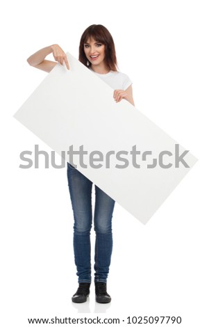 Young woman is standing, holding white placard, pointing at it and talking. Full length studio shot isolated on white.
